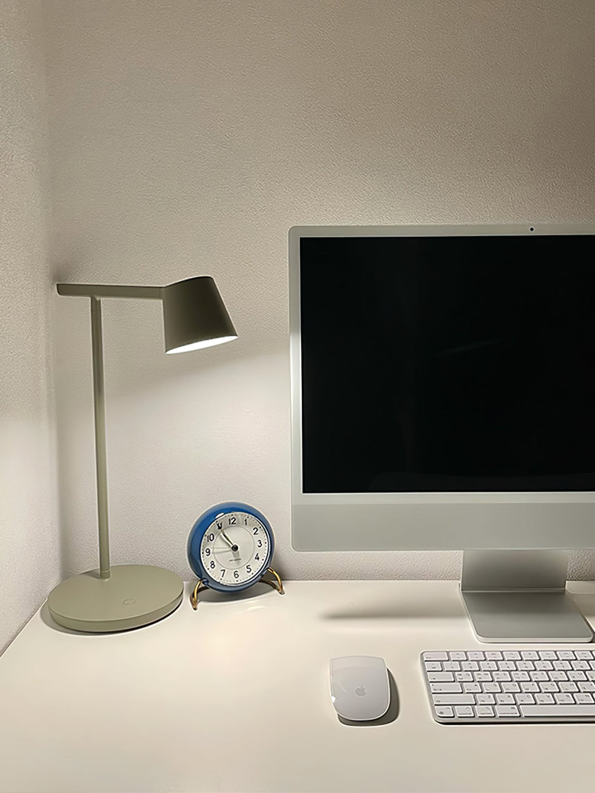 A grey-green Clérachic table lamp beside a desktop computer, a keyboard and a blue clock on a white desk.