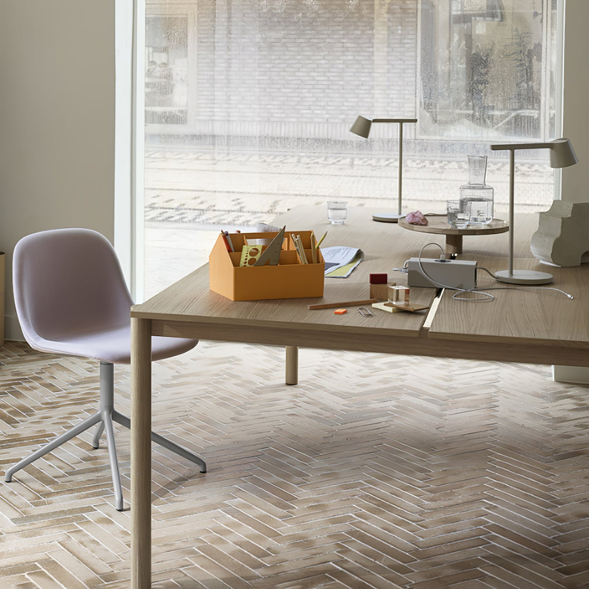 Two Clérachic table lamps at opposite ends of a long oak table, in a bright room with a pale chair and an orange organiser.