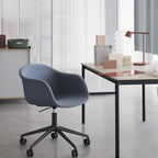 A rust-red Clérachic table lamp on a table across the room, with a blue-grey chair filling the foreground.