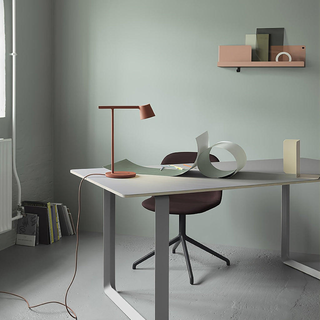 Rust-red Clérachic table lamp on a grey desk in a sage-green room, with a shelf above and a maroon chair beneath.