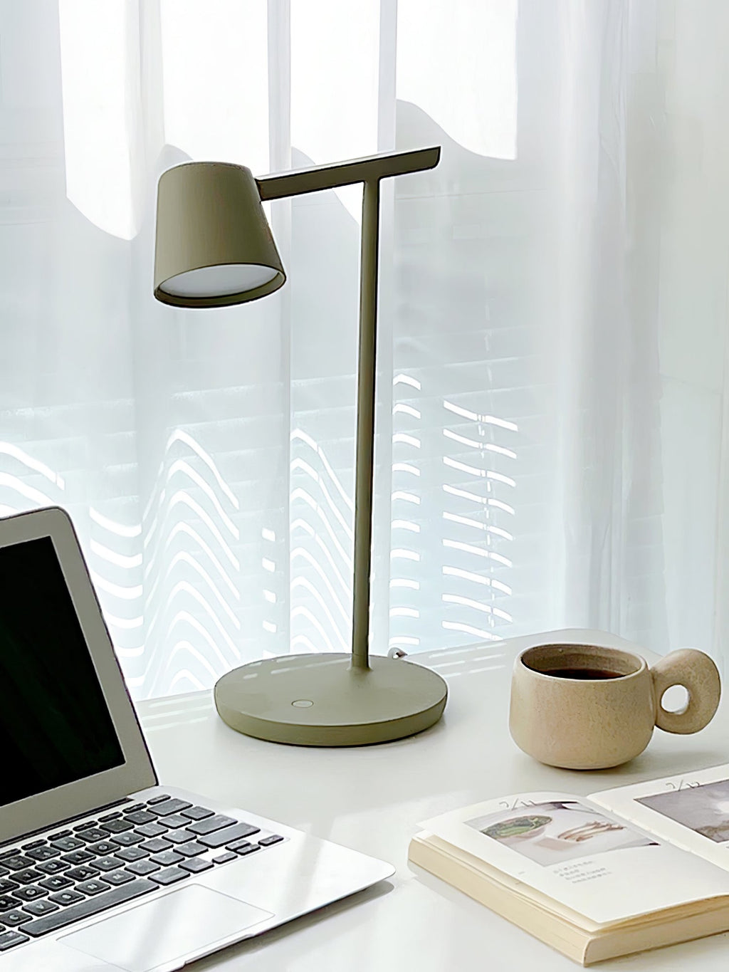 This Clérachic table lamp in olive switched on beside a laptop, a stoneware cup and an open book.