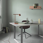 A rust-red Clérachic table lamp on a grey-topped desk, with a long cord trailing across the floor and a curled sheet beside it.