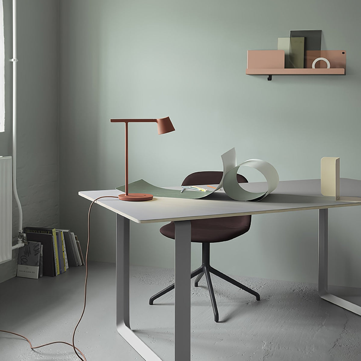A rust-red Clérachic table lamp on a grey-topped desk, with a long cord trailing across the floor and a curled sheet beside it.