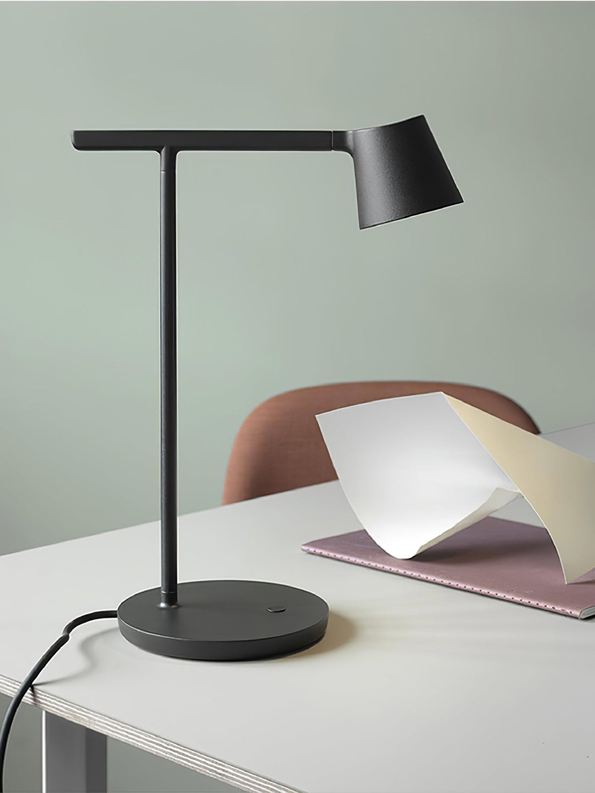 Black Clérachic table lamp in close view on a white desk, with a sage-green wall and the edge of a pink notebook.