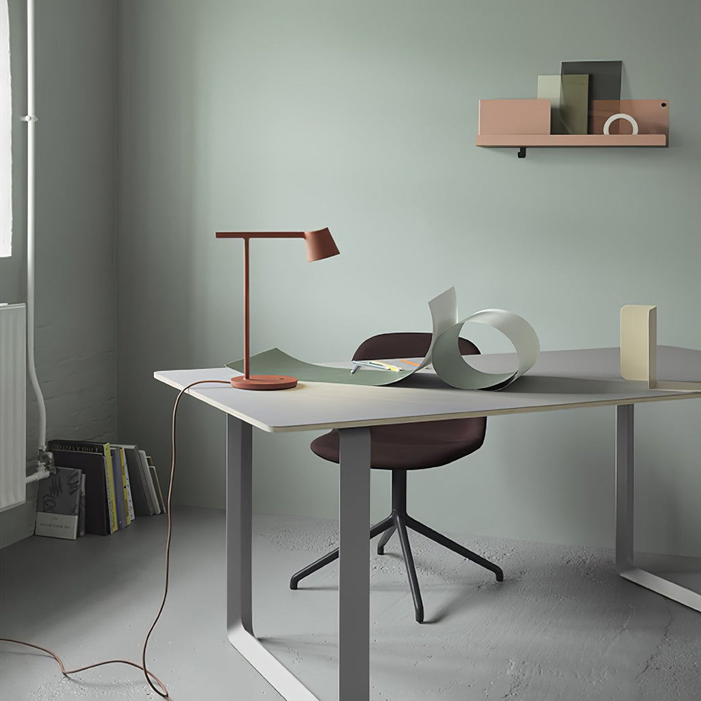 A rust-red Clérachic table lamp on a desk in a sage room, seen from further back, with books stacked on the floor.