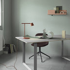 A rust-red Clérachic table lamp on a desk in a sage room, seen from further back, with books stacked on the floor.