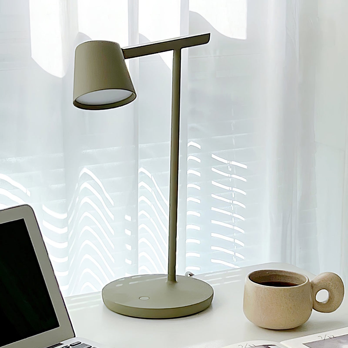 Close view of the olive head of a Clérachic table lamp, lit on the underside, with a laptop and a mug at the edges.