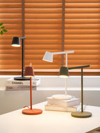 Four Clérachic table lamps in black, white, red and olive on a white desk in front of orange window blinds.