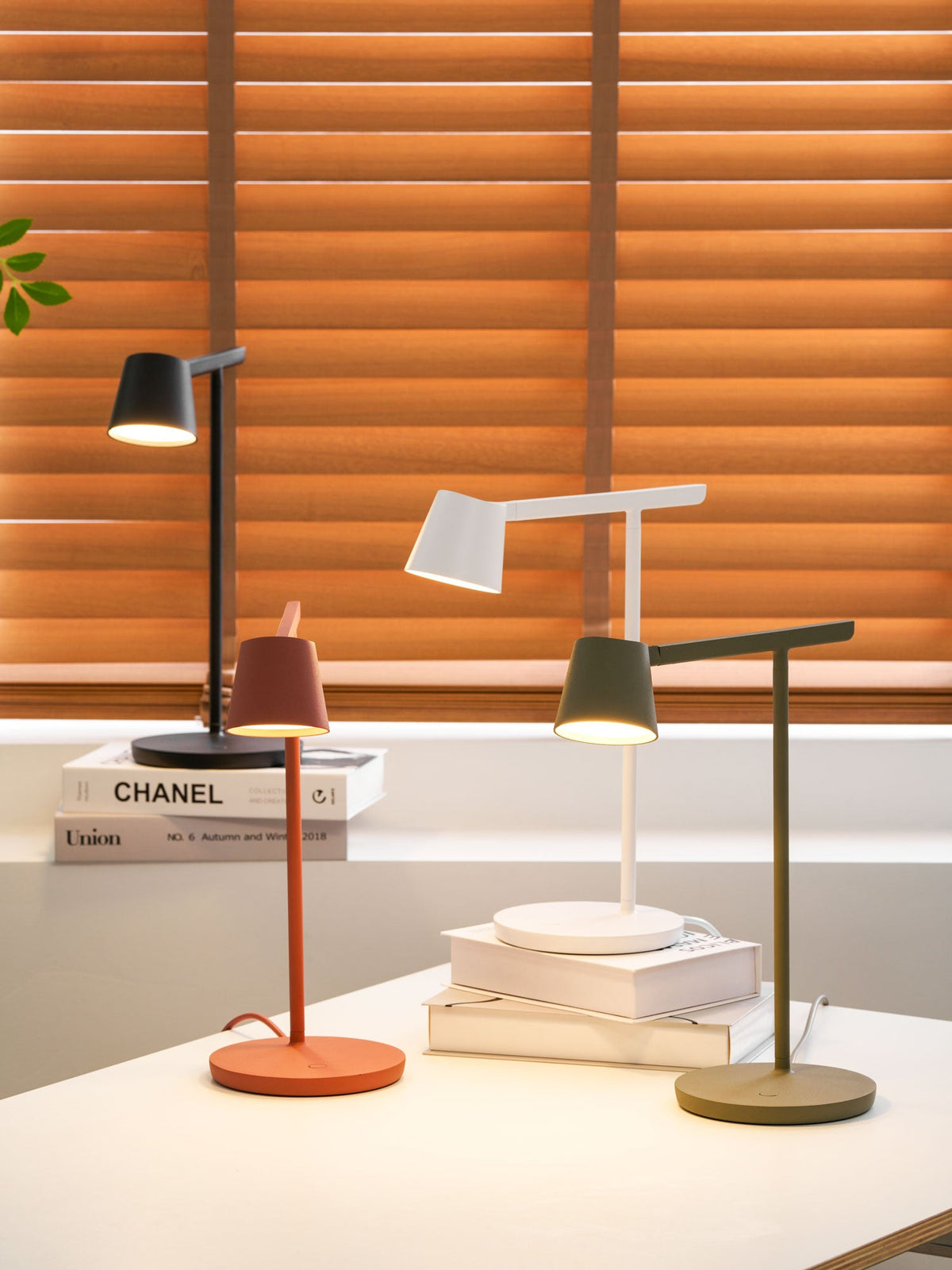 Four Clérachic table lamps in black, white, red and olive on a white desk in front of orange window blinds.