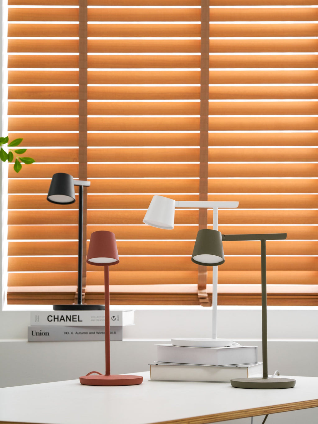 A wider view of four Clérachic table lamps grouped on a white desk, with the window frame and a leaf showing at the left.