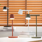 A Clérachic table lamp group with the red and olive heads switched on, set against orange blinds.