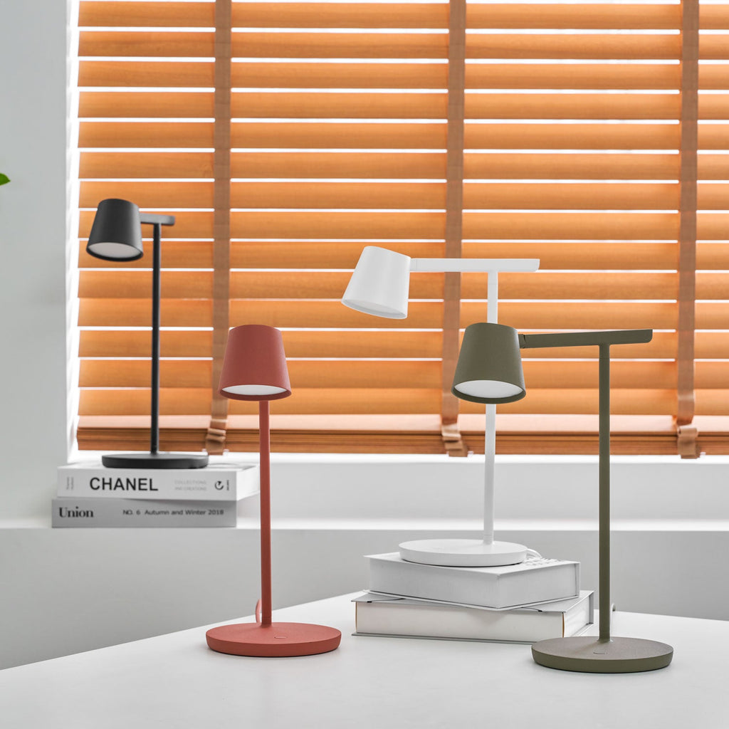 Four Clérachic table lamps on a white desk with the red and olive lamps lit and the blinds glowing orange behind.
