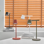 Four Clérachic table lamps on a white desk with the red and olive lamps lit and the blinds glowing orange behind.