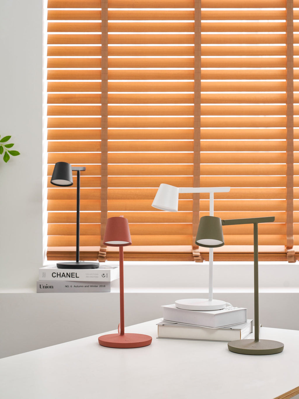 Four Clérachic table lamps from a distance, shown small in front of orange blinds on a bright white desk.