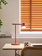 Tall view of a red Clérachic table lamp lit on a small table, with a checked mug and a cane chair at the bottom.