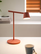 Red Clérachic table lamp lit, with a round base and a grey cup out of focus in the front, and a window behind.