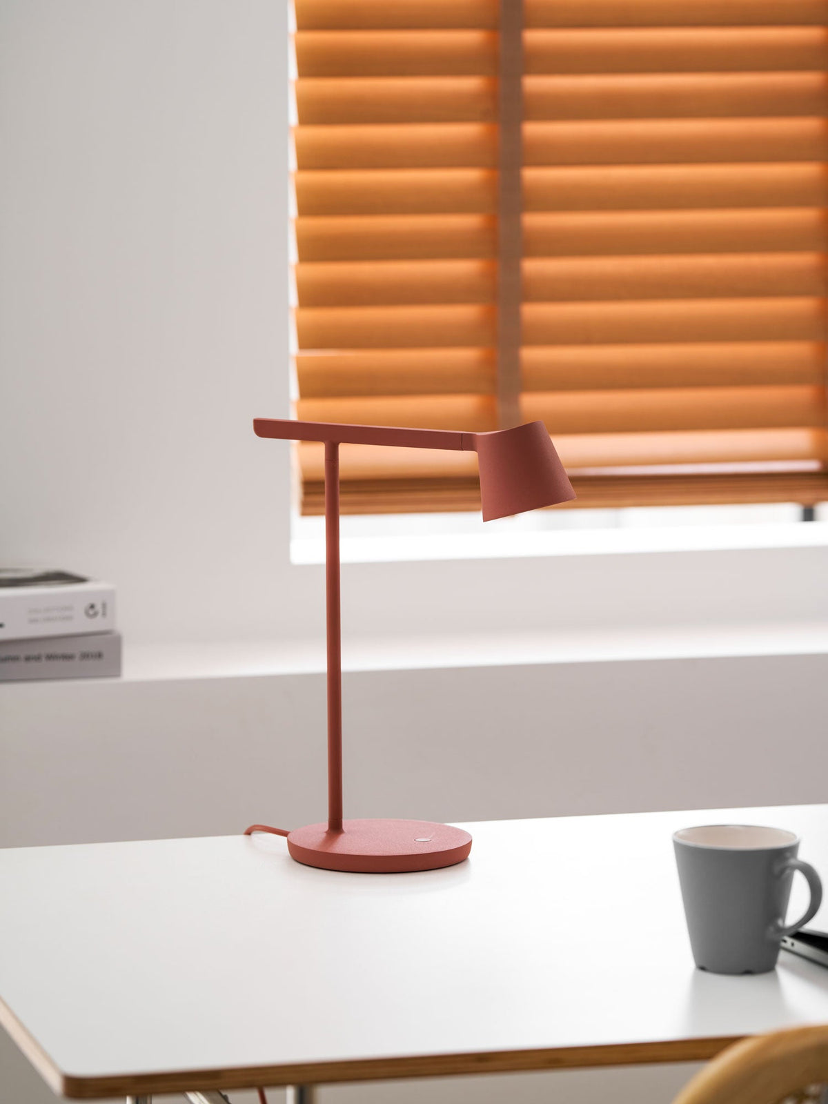 A red Clérachic table lamp on a white table with a grey mug beside it, the orange blinds blurred behind.
