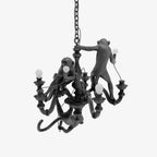 Black resin chandelier with three monkeys climbing its turned stem and candle-style arms, hung on a chain against a white backdrop, from Clérachic.