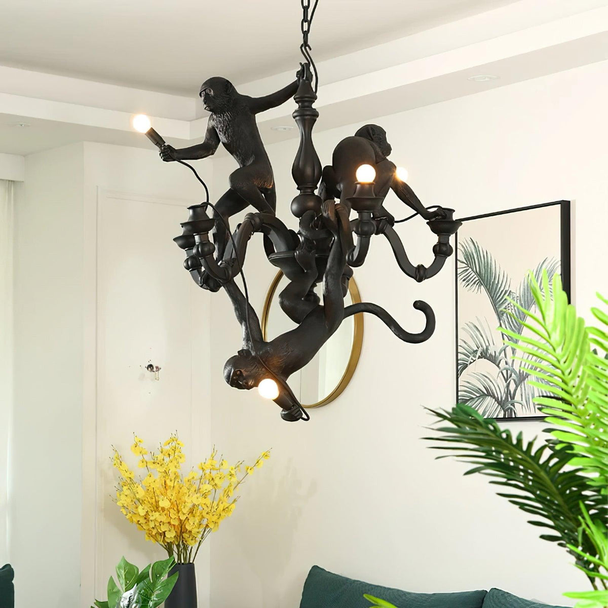 This Clérachic monkey chandelier in black, hung in a bright room with a gold-framed round mirror and green palm leaves behind.