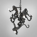 Black Clérachic chandelier with its three resin monkeys, one hanging by its tail with a bulb, on a plain grey background.