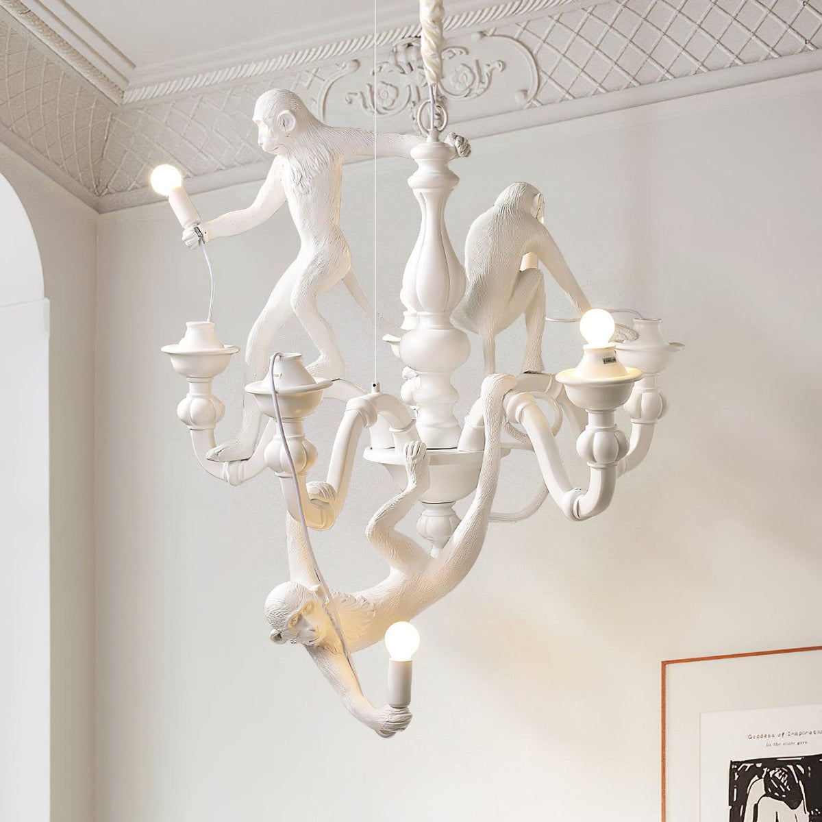 White Clérachic monkey chandelier hung from a ceiling rose in a pale room with decorative cornice moulding and a framed print.