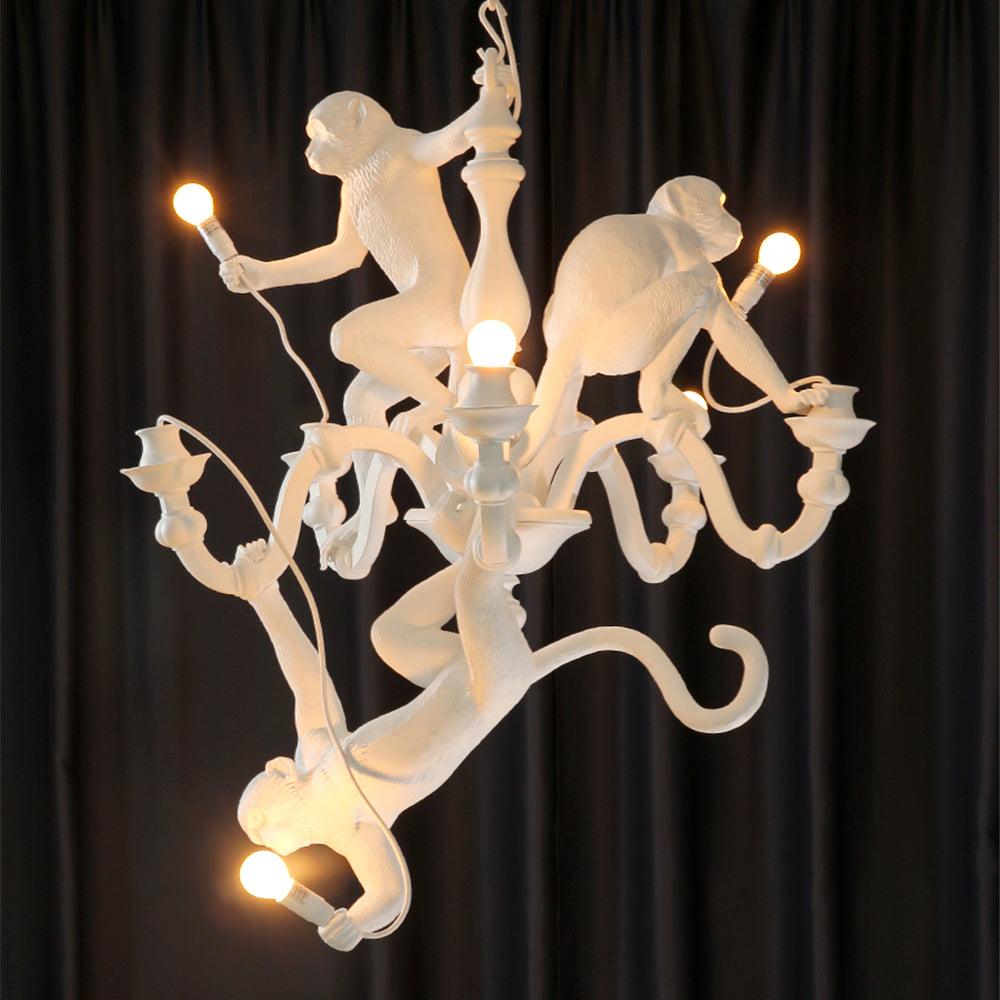 White Clérachic chandelier with lit bulbs glowing warmly against a dark curtain, one monkey hanging head-down from the lower arms.