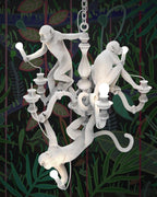 White Clérachic monkey chandelier lit in front of a dark botanical-print wall, one monkey holding a bulb at the top of the stem.