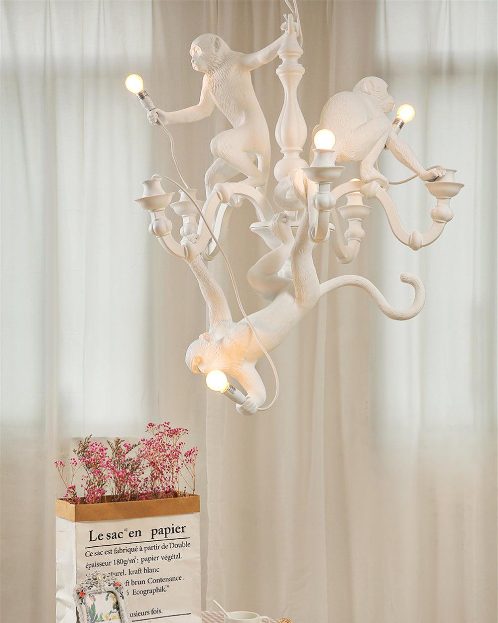 A Clérachic white monkey chandelier lit before sheer cream curtains, with a bag of pink flowers on a table beneath it.