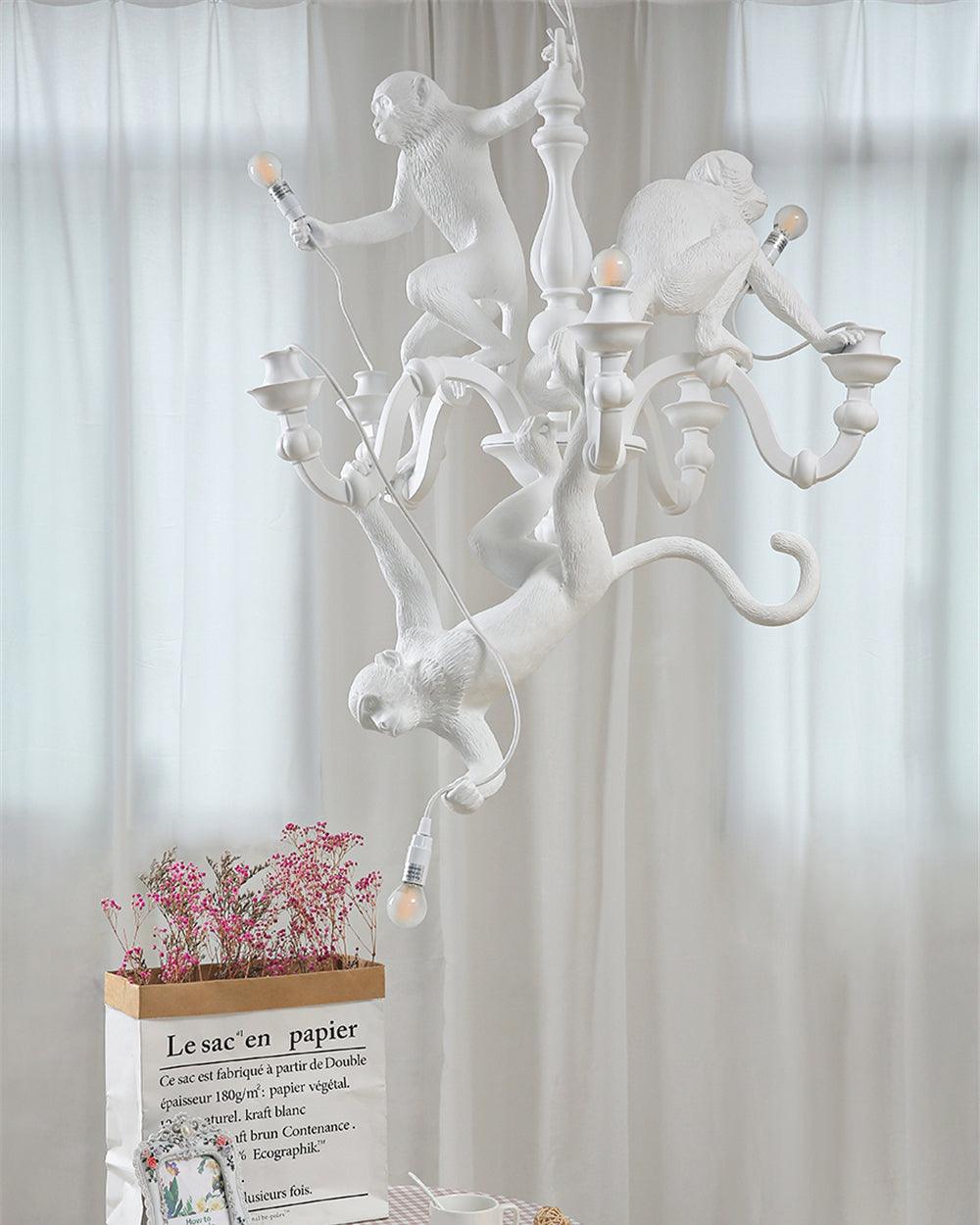 Unlit white Clérachic monkey chandelier hung in front of sheer curtains, with pink flowers in a paper bag below.