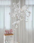Unlit white Clérachic monkey chandelier hung in front of sheer curtains, with pink flowers in a paper bag below.