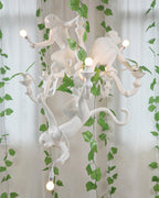 White Clérachic monkey chandelier lit among trailing green ivy in front of sheer curtains, bulbs shining from its candle-style arms.