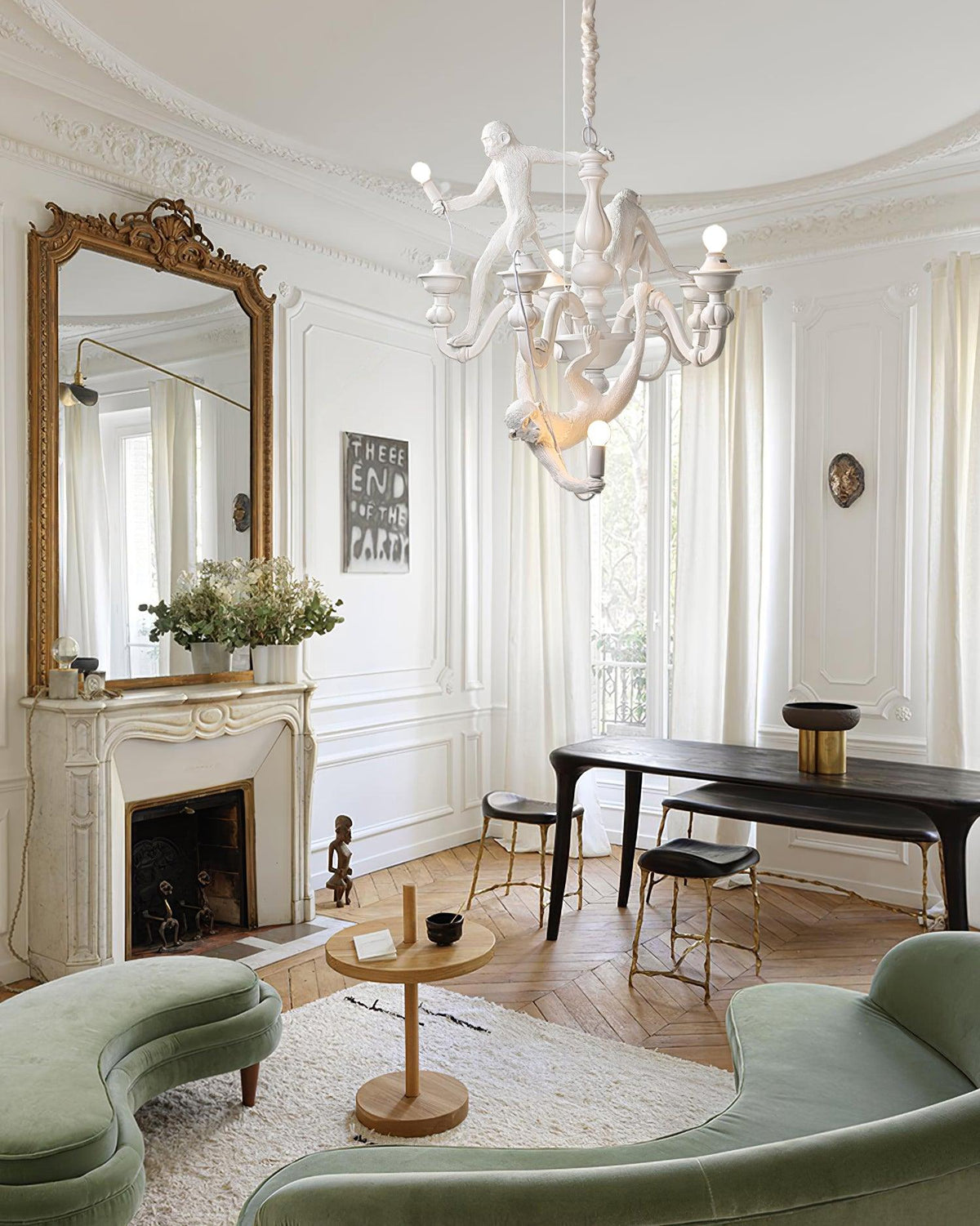 A Clérachic white chandelier hung in a tall Parisian-style room with a gilt mirror, a fireplace, a dark table and green velvet seating.