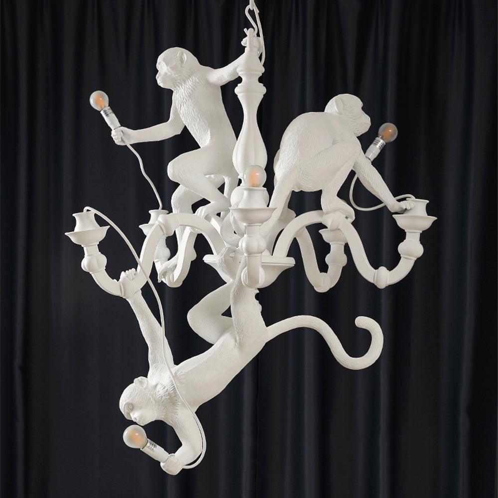 White Clérachic chandelier lit against dark curtains, three resin monkeys climbing its stem with a bulb in each hand or foot.