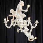 Close view of the three white monkeys on this Clérachic chandelier, each holding a lit bulb, against a dark navy curtain.
