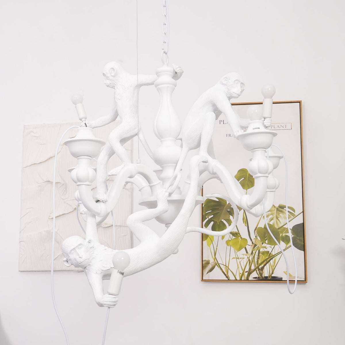 White Clérachic monkey chandelier beside a framed leaf print on a pale wall, one monkey hanging low by the arms.