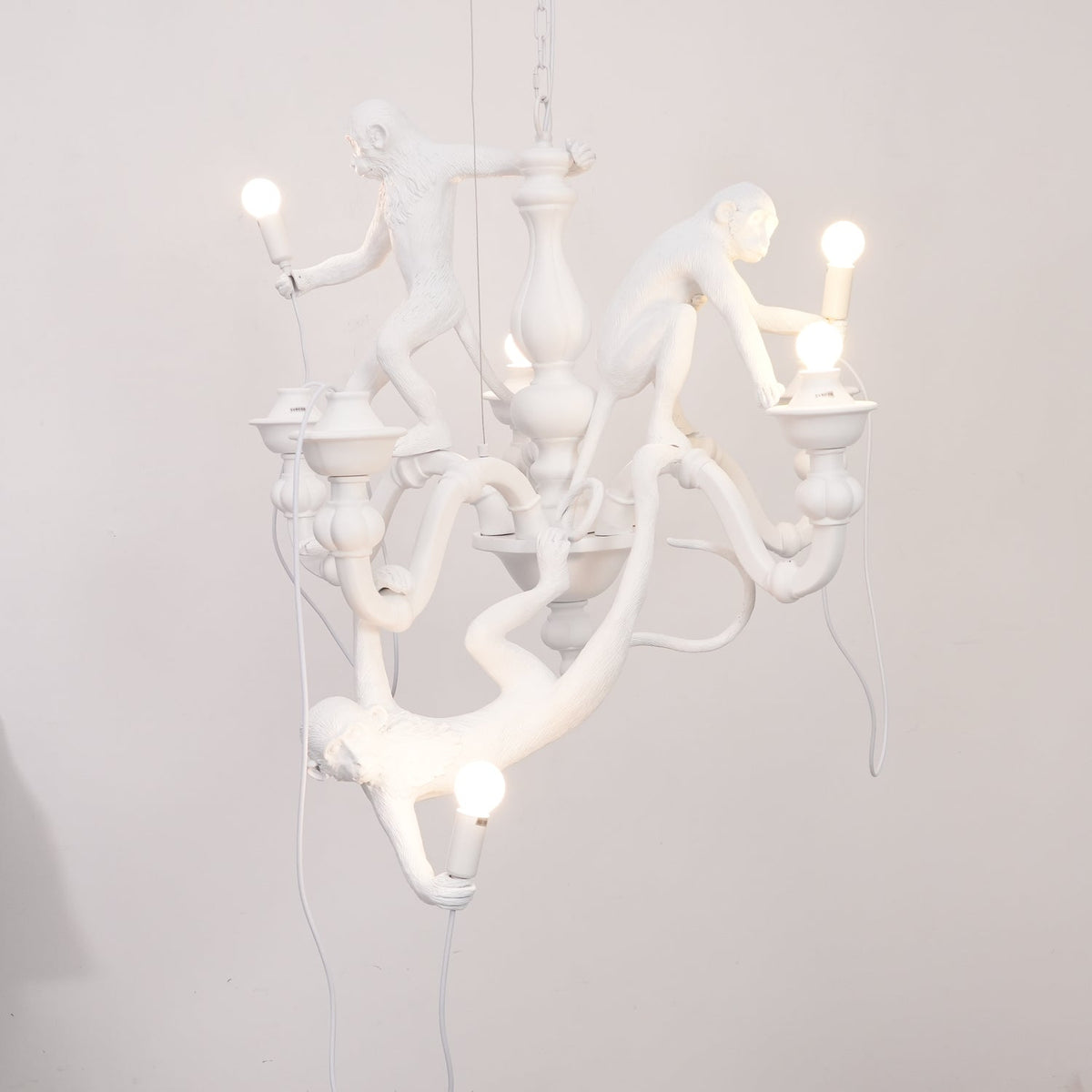 Lit white Clérachic monkey chandelier hung in front of a pale wall, with one monkey hanging below holding a glowing bulb.