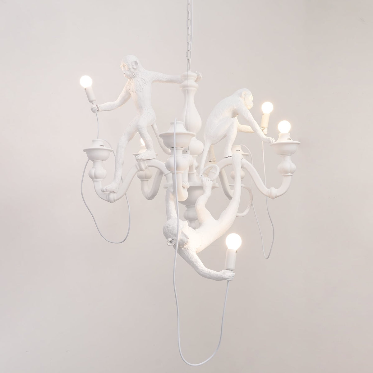 White Clérachic chandelier with the monkeys' bulbs lit, hung in front of a plain pale wall and trailing a white cable.