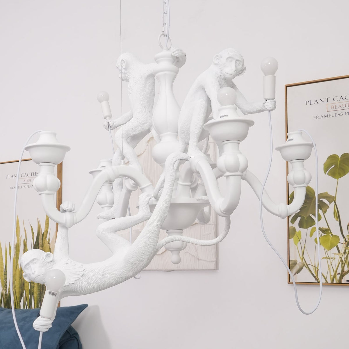 Side view of this white Clérachic monkey chandelier against a pale wall, with a framed botanical print and a blue cushion behind.