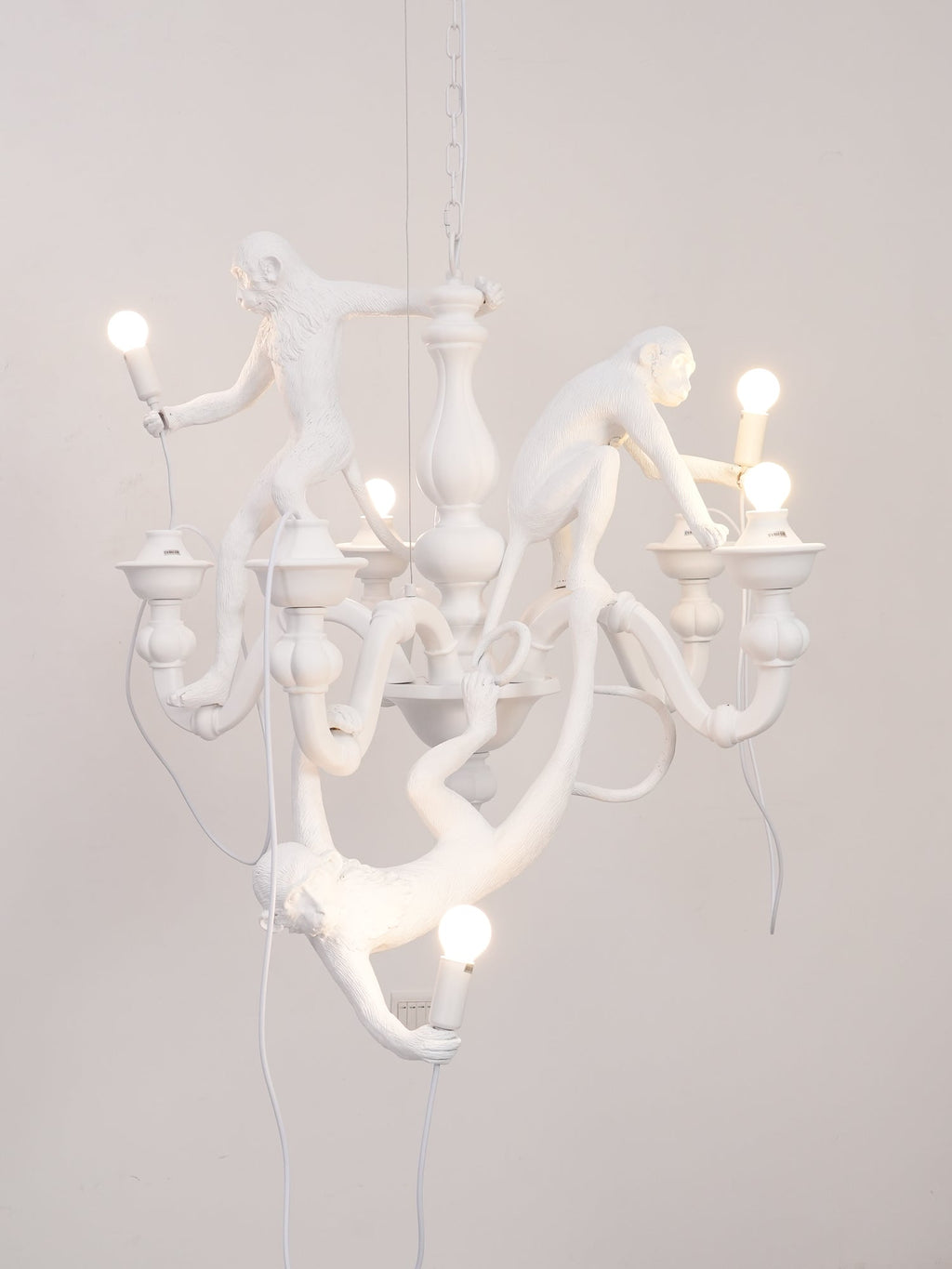 White Clérachic monkey chandelier lit against a soft pink-grey wall, its resin monkeys reaching toward the bulbs.