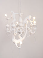 White Clérachic monkey chandelier lit against a soft pink-grey wall, its resin monkeys reaching toward the bulbs.