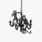 Black Clérachic monkey chandelier with its resin monkeys and lit bulbs, hung on a chain against a light grey background.