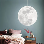 Clérachic large full-moon pendant glowing white beside a bed with blush bedding, against a muted blue-green wall.