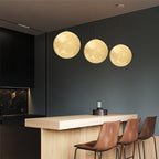Clérachic three moon pendants at graduated heights above a kitchen bar, glowing warm against a dark green wall.