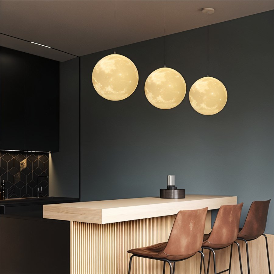 Clérachic three moon pendants at graduated heights above a kitchen bar, glowing warm against a dark green wall.