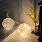 Two lit moon spheres from Clérachic sit on a glossy floor beside a glass vase of yellow flowers in a dim corner.