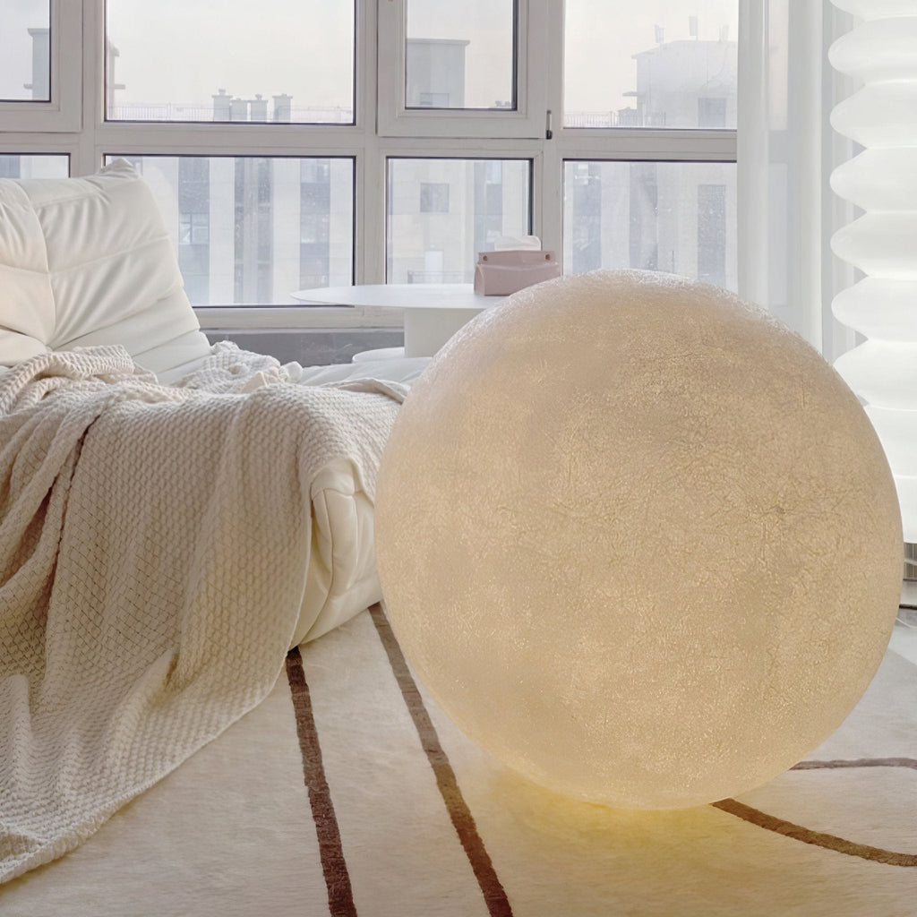 A large Clérachic moon lamp glows softly on a striped cream rug beside a bed with a knitted throw, in front of a tall window.