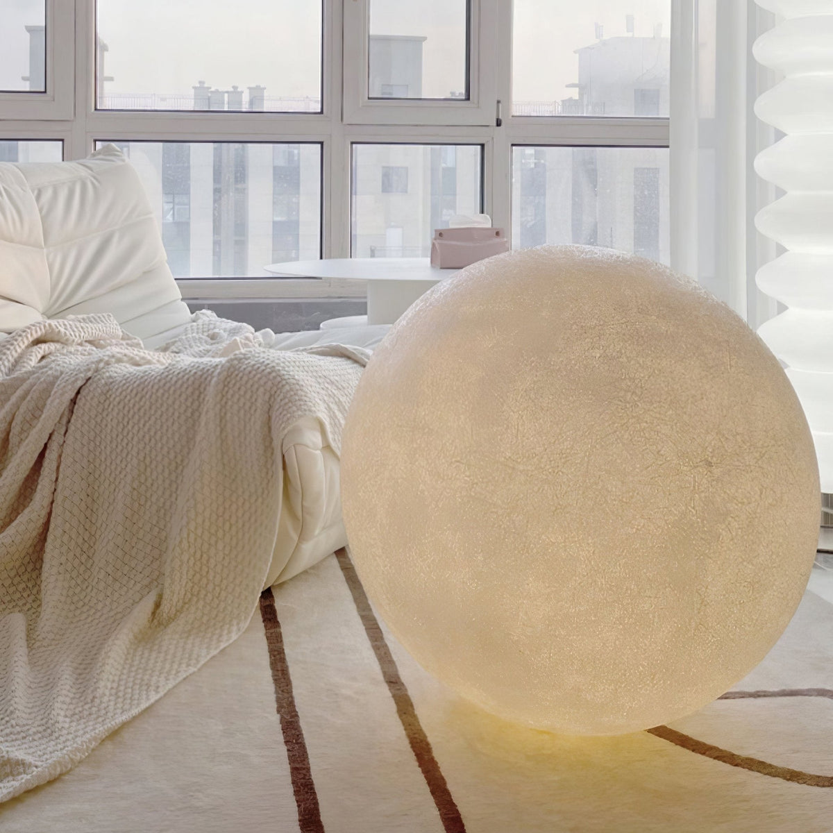 A large Clérachic moon lamp glows softly on a striped cream rug beside a bed with a knitted throw, in front of a tall window.