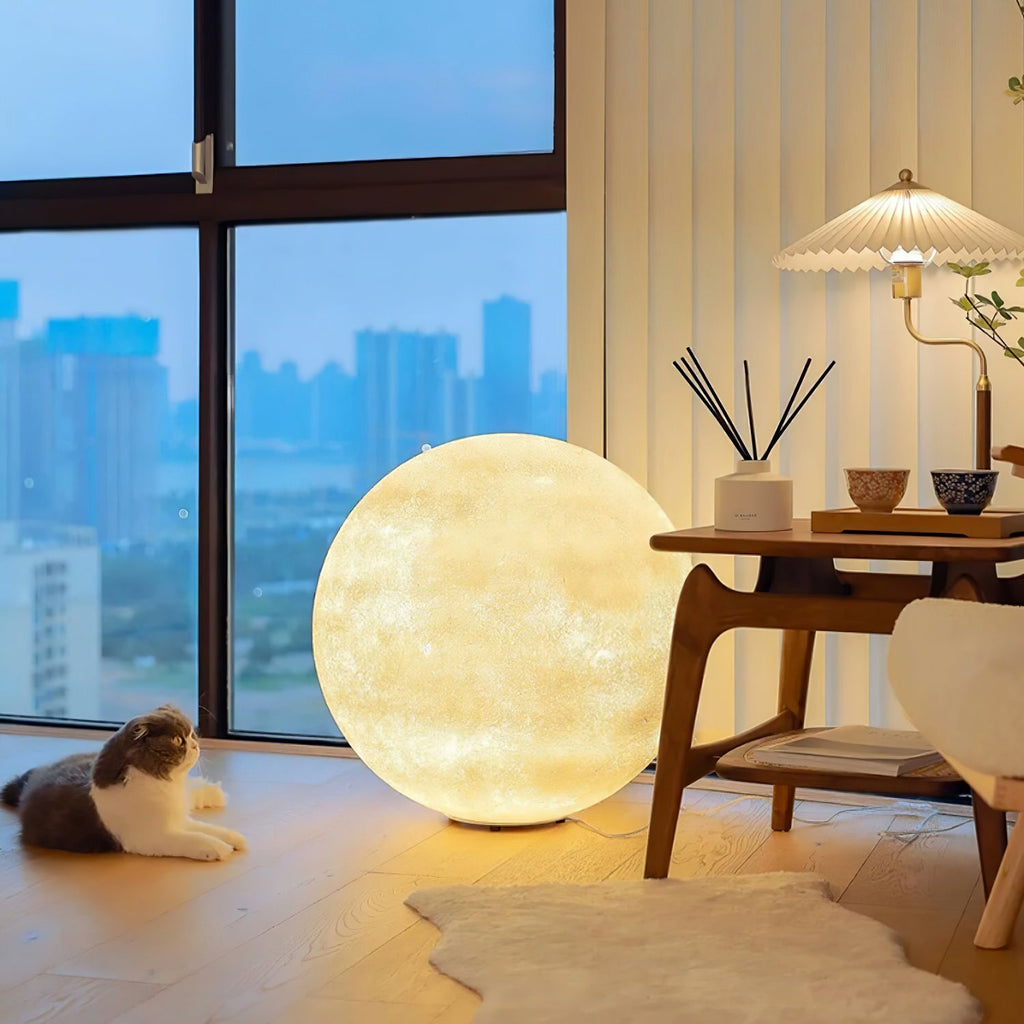 A warm-glowing Clérachic moon lamp stands by a window with a city view, a wooden side table, a cat and a white fur rug.