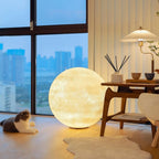 A warm-glowing Clérachic moon lamp stands by a window with a city view, a wooden side table, a cat and a white fur rug.