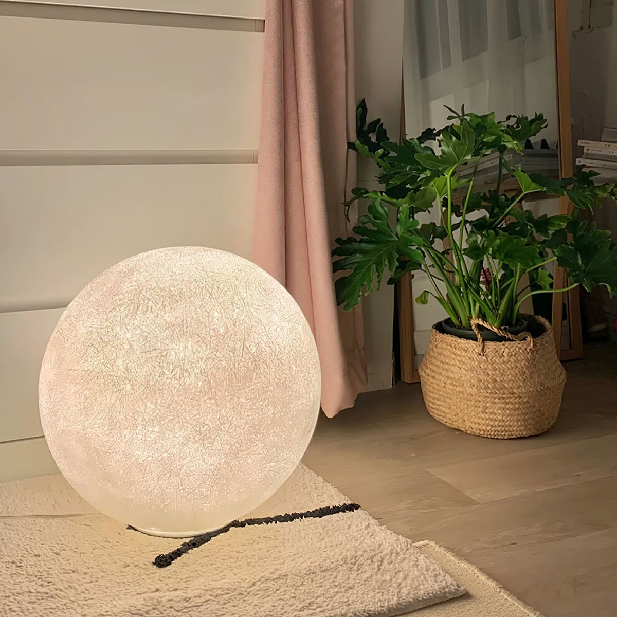 The Clérachic moon lamp sits on a cream rug against a wall, next to a leafy plant in a woven basket and a pink curtain.
